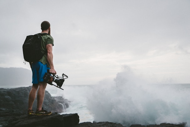 A person with a backpack stands on rocky terrain next to the ocean, holding a camera rig while looking at crashing waves. The sky is cloudy, creating a dramatic and adventurous atmosphere.