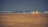 Two lifeguards wearing yellow jackets and orange shorts stand on a sandy beach, facing the sea with rescue equipment at their feet. In the distance, two people are sitting near the shoreline, observing the waves. The sky is overcast, creating a moody atmosphere.