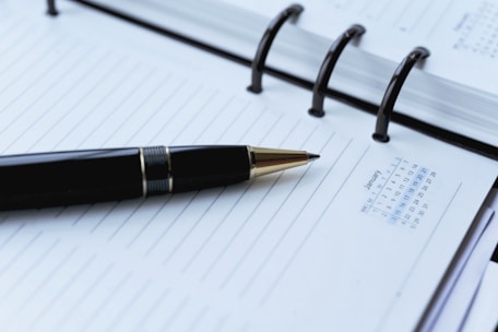 A black pen is resting on an open spiral-bound planner or notebook with lined pages. The planner shows a small monthly calendar for January on the right side of the page. The focus is on the pen and the planner's neat, organized layout.