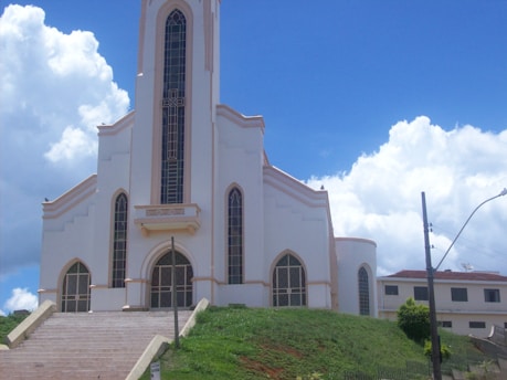 A large, white church building with tall windows and a central tower is positioned on a grassy hill. The sky is bright blue with scattered white clouds. Steps lead up to the entrance, and there is a lamp post and some greenery in the foreground.