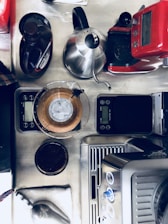 A countertop set up for coffee making, featuring a variety of coffee equipment including a red coffee machine, a stainless steel kettle, a digital scale, a pour-over coffee maker with a paper filter visible, and a container of ground coffee. The arrangement is organized, suggesting a functional and ready-to-use coffee station.