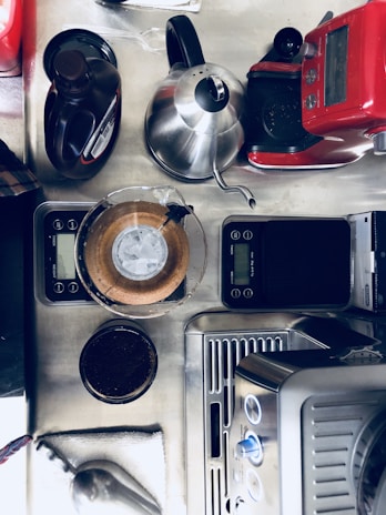 A countertop set up for coffee making, featuring a variety of coffee equipment including a red coffee machine, a stainless steel kettle, a digital scale, a pour-over coffee maker with a paper filter visible, and a container of ground coffee. The arrangement is organized, suggesting a functional and ready-to-use coffee station.