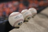 A row of official Major League Baseballs resting on a concrete surface, with the focus on the baseball closest to the viewer. The background is blurred, featuring a hint of orange color, possibly from a crowd or seats.