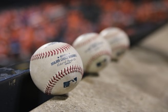 A row of official Major League Baseballs resting on a concrete surface, with the focus on the baseball closest to the viewer. The background is blurred, featuring a hint of orange color, possibly from a crowd or seats.