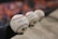 A row of official Major League Baseballs resting on a concrete surface, with the focus on the baseball closest to the viewer. The background is blurred, featuring a hint of orange color, possibly from a crowd or seats.