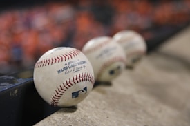 A row of official Major League Baseballs resting on a concrete surface, with the focus on the baseball closest to the viewer. The background is blurred, featuring a hint of orange color, possibly from a crowd or seats.