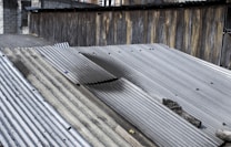 A series of corrugated metal sheets form a rooftop with visible aging and weathering. The metal sheets have patches of rust and discoloration, and the edges appear worn. In the background, a wooden fence adds texture with its vertical planks and varying shades of brown. Some debris, possibly rocks or dirt, is scattered on the rooftop.