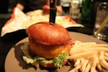 A burger with a knife lodged into the top bun is served on a plate, accompanied by crispy golden French fries. The bun is glossy, the cheese is melted, and there are fresh lettuce leaves visible beneath the patty.