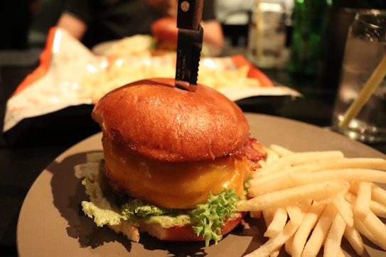 A burger with a knife lodged into the top bun is served on a plate, accompanied by crispy golden French fries. The bun is glossy, the cheese is melted, and there are fresh lettuce leaves visible beneath the patty.