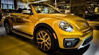 A bright yellow convertible car is parked indoors, showcasing its sleek design and modern features. The headlights are prominent, and the car has distinctive black accents and stylish alloy wheels. Reflections on the car's surface suggest a well-lit showroom environment with other vehicles visible in the background.
