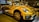 A bright yellow convertible car is parked indoors, showcasing its sleek design and modern features. The headlights are prominent, and the car has distinctive black accents and stylish alloy wheels. Reflections on the car's surface suggest a well-lit showroom environment with other vehicles visible in the background.