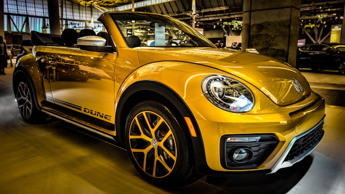 A bright yellow convertible car is parked indoors, showcasing its sleek design and modern features. The headlights are prominent, and the car has distinctive black accents and stylish alloy wheels. Reflections on the car's surface suggest a well-lit showroom environment with other vehicles visible in the background.