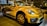 A bright yellow convertible car is parked indoors, showcasing its sleek design and modern features. The headlights are prominent, and the car has distinctive black accents and stylish alloy wheels. Reflections on the car's surface suggest a well-lit showroom environment with other vehicles visible in the background.