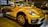 A bright yellow convertible car is parked indoors, showcasing its sleek design and modern features. The headlights are prominent, and the car has distinctive black accents and stylish alloy wheels. Reflections on the car's surface suggest a well-lit showroom environment with other vehicles visible in the background.