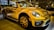 A bright yellow convertible car is parked indoors, showcasing its sleek design and modern features. The headlights are prominent, and the car has distinctive black accents and stylish alloy wheels. Reflections on the car's surface suggest a well-lit showroom environment with other vehicles visible in the background.