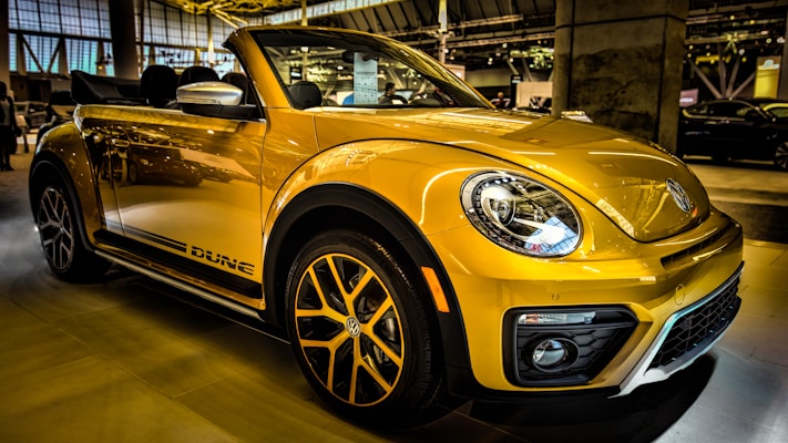 A bright yellow convertible car is parked indoors, showcasing its sleek design and modern features. The headlights are prominent, and the car has distinctive black accents and stylish alloy wheels. Reflections on the car's surface suggest a well-lit showroom environment with other vehicles visible in the background.