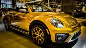 A bright yellow convertible car is parked indoors, showcasing its sleek design and modern features. The headlights are prominent, and the car has distinctive black accents and stylish alloy wheels. Reflections on the car's surface suggest a well-lit showroom environment with other vehicles visible in the background.