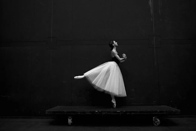 A ballet dancer balances gracefully on pointe with one leg extended behind her. She wears a flowing tutu and a fitted bodice against a dark, minimalist backdrop.