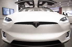 A sleek white car with distinctive gull-wing doors is prominently displayed in a modern showroom. The car features a prominent logo on the front and has a futuristic design with bright headlights. The background includes digital screens and a few silhouetted figures.