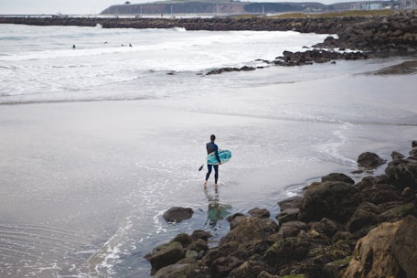 A surfer wearing a sleek wetsuit walking along the shoreline with waves in the background.