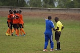 A group of soccer players wearing orange jerseys and shorts are engaged in a group hug, celebrating on a grassy field. In the foreground, a coach in a blue tracksuit is pointing while talking to a referee in a yellow shirt and black shorts.