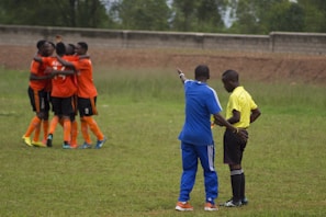 A group of soccer players wearing orange jerseys and shorts are engaged in a group hug, celebrating on a grassy field. In the foreground, a coach in a blue tracksuit is pointing while talking to a referee in a yellow shirt and black shorts.