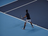 A tennis player is on a blue hard court, preparing to hit a backhand shot. The player is wearing a black shirt, white shorts, and sneakers. The court is marked with white lines, and the player is holding a tennis racket with both hands.