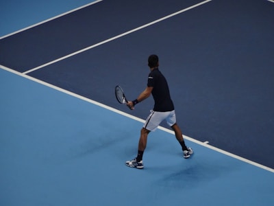 A tennis player is on a blue hard court, preparing to hit a backhand shot. The player is wearing a black shirt, white shorts, and sneakers. The court is marked with white lines, and the player is holding a tennis racket with both hands.