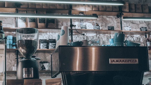 A cozy coffee shop setting displays a modern espresso machine with the brand name La Marzocco prominently on the front. A coffee grinder labeled Compak filled with coffee beans is positioned nearby. Several blue cups are stacked on the espresso machine, and numerous paper cups with a logo sit adjacent. Shelves above contain assorted coffee bags and merchandise under soft fluorescent lighting.