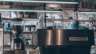A cozy coffee shop setting displays a modern espresso machine with the brand name La Marzocco prominently on the front. A coffee grinder labeled Compak filled with coffee beans is positioned nearby. Several blue cups are stacked on the espresso machine, and numerous paper cups with a logo sit adjacent. Shelves above contain assorted coffee bags and merchandise under soft fluorescent lighting.
