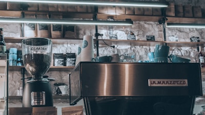 A cozy coffee shop setting displays a modern espresso machine with the brand name La Marzocco prominently on the front. A coffee grinder labeled Compak filled with coffee beans is positioned nearby. Several blue cups are stacked on the espresso machine, and numerous paper cups with a logo sit adjacent. Shelves above contain assorted coffee bags and merchandise under soft fluorescent lighting.