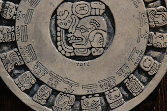 A circular stone carving featuring intricate glyphs and symbols, characteristic of ancient Mesoamerican or Mayan art. The central portion showcases a prominent face surrounded by various other detailed illustrations, arranged like a calendar.