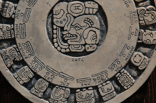 A circular stone carving featuring intricate glyphs and symbols, characteristic of ancient Mesoamerican or Mayan art. The central portion showcases a prominent face surrounded by various other detailed illustrations, arranged like a calendar.