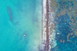 An aerial view of a coral reef and rocky barrier separating blue-green ocean waters. Two small structures, possibly platforms, are visible in the water on either side of the reef.