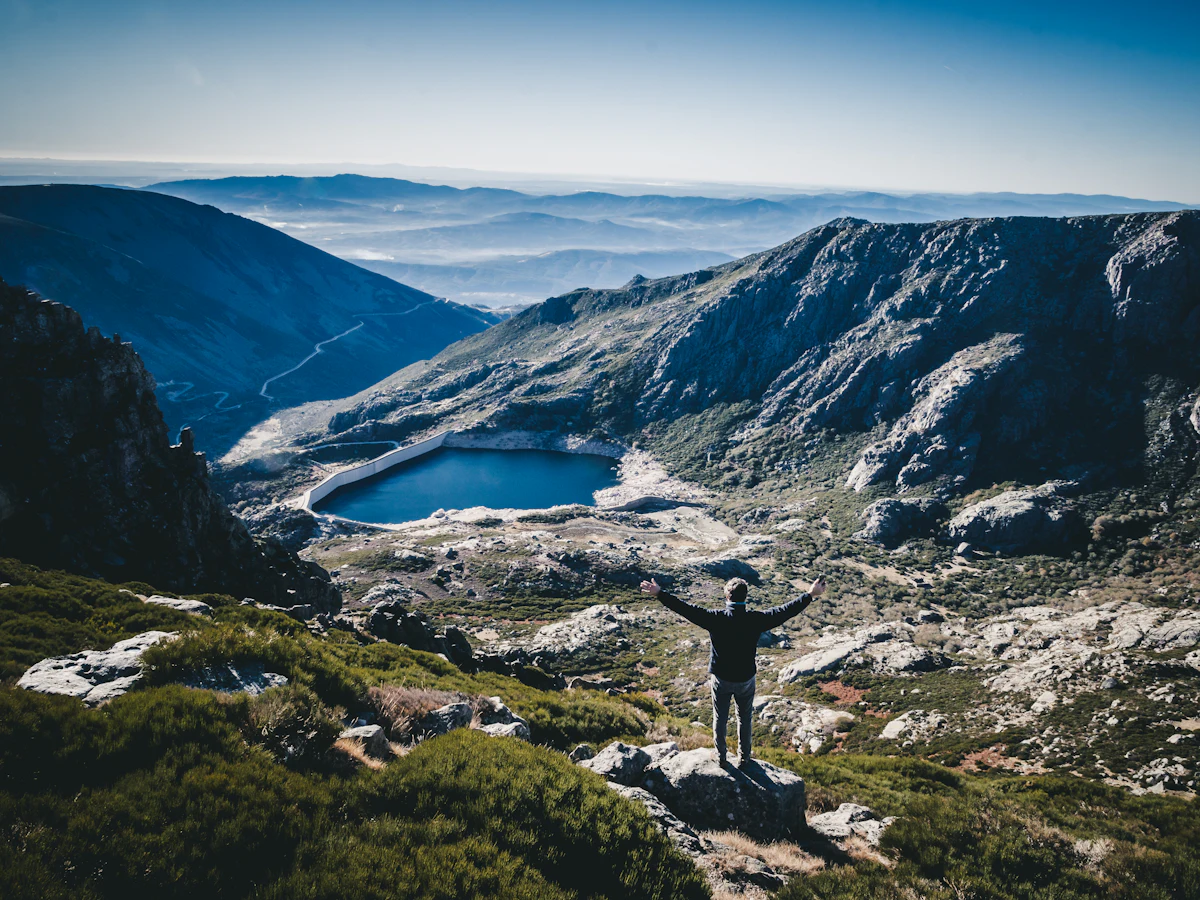 A hiker with arms raised on a boulder above a lake in a rocky valley of the Serra da Estrela