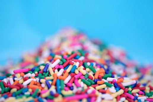 A pile of colorful candy sprinkles scattered across a surface with a blurred background. The sprinkles are in various bright colors including pink, green, yellow, blue, orange, and purple.