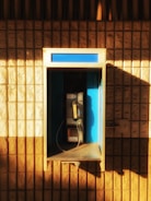 A vintage payphone is mounted on a wall with textured brick, partially illuminated by warm sunlight. The phone booth features a blue interior with metallic components and a yellow handset.