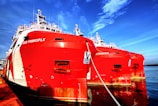 Two large red ships are docked side by side in a harbor, showcasing intricate structures and equipment on their decks. The vibrant red hulls of the vessels contrast sharply with the blue sea and sky in the background.