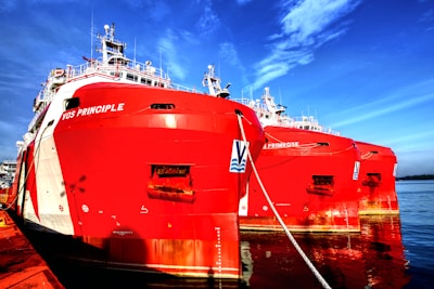 Two large red ships are docked side by side in a harbor, showcasing intricate structures and equipment on their decks. The vibrant red hulls of the vessels contrast sharply with the blue sea and sky in the background.