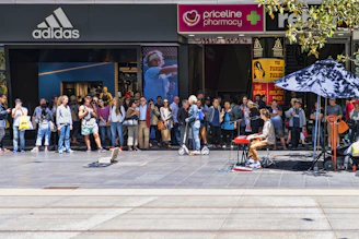 A street performer playing a keyboard entertains a crowd gathered in front of several retail stores, including Adidas and Priceline Pharmacy. The audience appears engaged, with some taking photos and others watching intently.