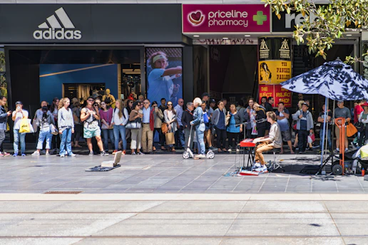 A street performer playing a keyboard entertains a crowd gathered in front of several retail stores, including Adidas and Priceline Pharmacy. The audience appears engaged, with some taking photos and others watching intently.