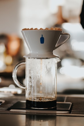 A coffee dripper placed on top of a glass carafe is brewing coffee. The dripper features a simple design with a blue bottle logo, and steam is visible within the carafe as the coffee drips in.