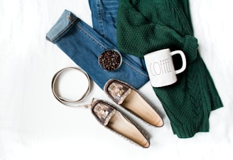 A neatly arranged flat lay featuring a pair of jeans, a green knitted sweater, a pair of patterned loafers, a metal belt, a mug labeled 'COFFEE', and a bowl of coffee beans. The items are placed on a white background, indicating a casual, cozy theme.
