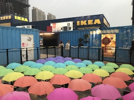 A display of numerous colorful umbrellas, arranged in rows, with an IKEA store and high-rise buildings visible in the background. Two people are standing near a blue shipping container decorated with plants and artwork, while festive decorations can be seen through a window.