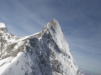 A steep, snow-covered mountain peak rises sharply against a clear blue sky. The rugged texture of the rocky slopes is visible, with areas of snow clinging to the surface. The sun casts shadows, accentuating the sharp angles and steepness of the peak.