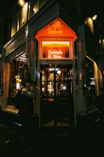 A cozy bar entrance is illuminated by a warm neon sign reading 'Cocktails & Bites'. The interior appears inviting with dim lighting and a visible bar stocked with glasses and bottles. The dark exterior contrasts with the vibrant glow of the sign, creating an intimate atmosphere. A person walks by, slightly obscured by shadows.