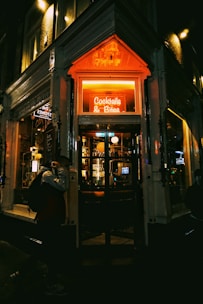 A cozy bar entrance is illuminated by a warm neon sign reading 'Cocktails & Bites'. The interior appears inviting with dim lighting and a visible bar stocked with glasses and bottles. The dark exterior contrasts with the vibrant glow of the sign, creating an intimate atmosphere. A person walks by, slightly obscured by shadows.