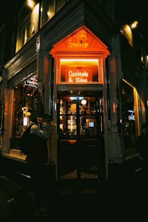A cozy bar entrance is illuminated by a warm neon sign reading 'Cocktails & Bites'. The interior appears inviting with dim lighting and a visible bar stocked with glasses and bottles. The dark exterior contrasts with the vibrant glow of the sign, creating an intimate atmosphere. A person walks by, slightly obscured by shadows.
