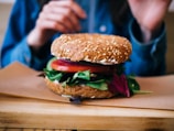 A close-up view of a multigrain sandwich with sesame seeds, filled with fresh vegetables including tomato slices and leafy greens. The sandwich is placed on a wooden board with hands partially visible in the background, suggesting a meal setting.