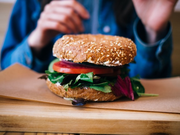 A close-up view of a multigrain sandwich with sesame seeds, filled with fresh vegetables including tomato slices and leafy greens. The sandwich is placed on a wooden board with hands partially visible in the background, suggesting a meal setting.